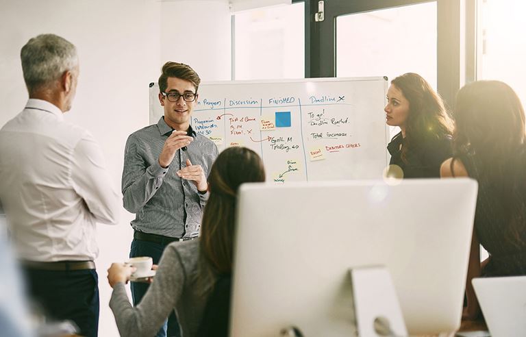 Multiple people listening to a man speak in front of a white board