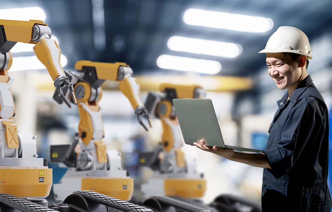 Factory worker holding a laptop computer while examining robotic equipment