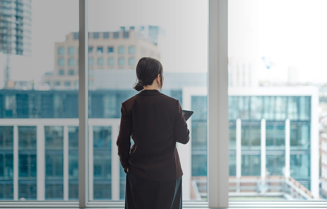 Young businesswoman with digital tablet, standing against office window with city view in modern commercial building.