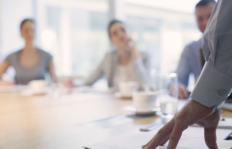 Focus is on a hand on a boardroom table with the other meeting attendees blurred in the background.