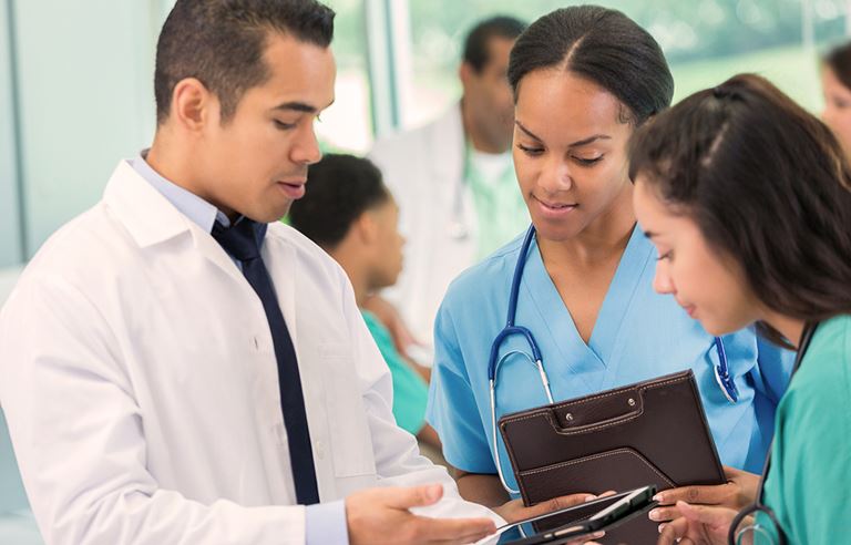 Image of two medical women in scrubs and a man in a white lab coat looking at a tablet screen.