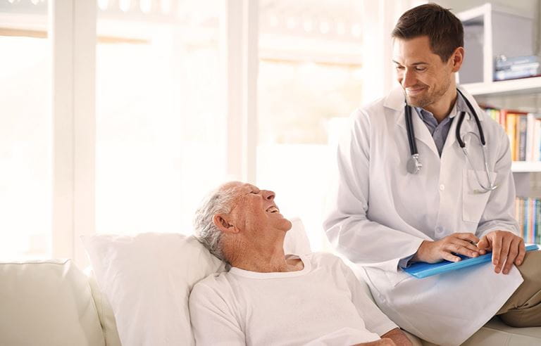 Image of elderly man in hospital bed with a male doctor sitting on the edge smiling down at him; SNF interrupted stay PDPM.