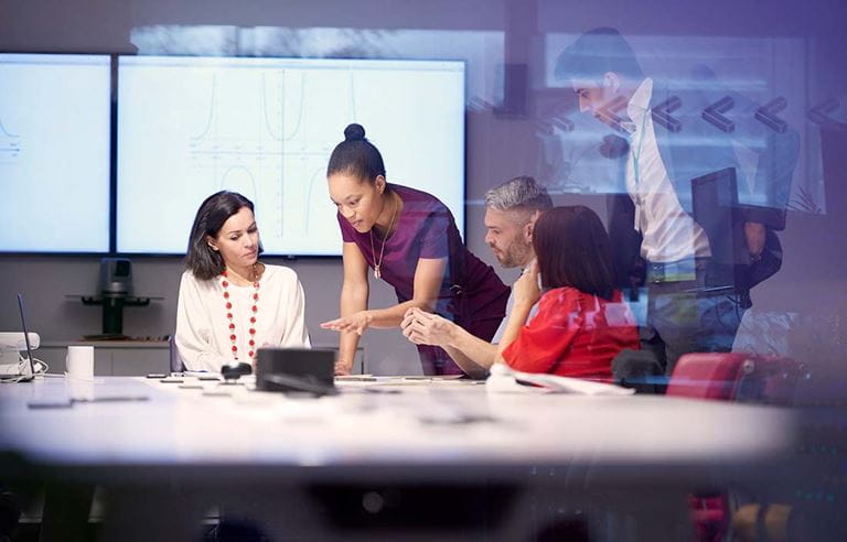 Image of a business meeting in a conference room with large screens in the background.