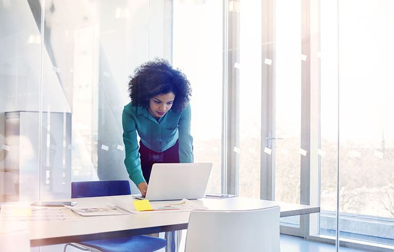 Younger businesswoman leaning over a table using a laptop computer.