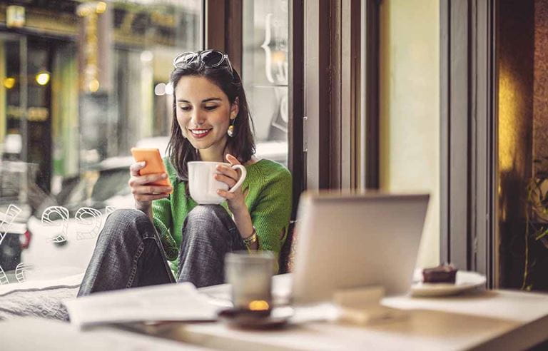 Woman sitting in a cafe holding a cup of coffee and using her phone.