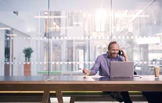Man using cell phone smiling while sitting at a table using his laptop computer.