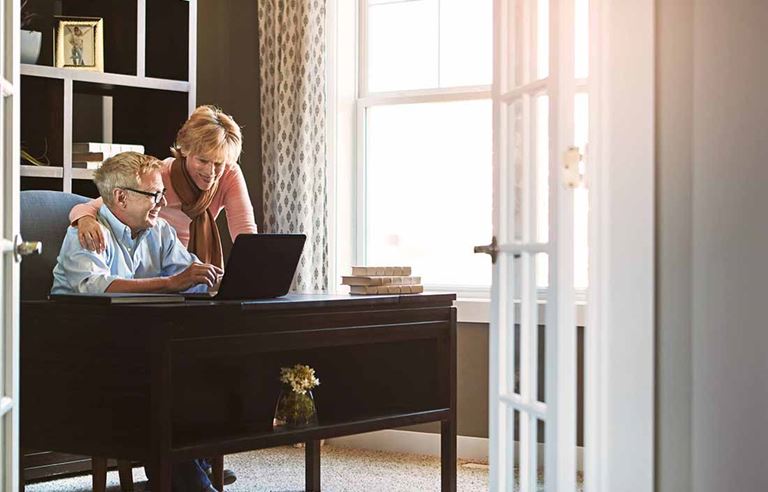 Two elderly people by a wooden desk using a laptop computer smiling.