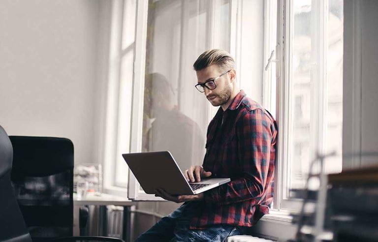 Younger business professional in casual clothing leaning against a window using a laptop computer.