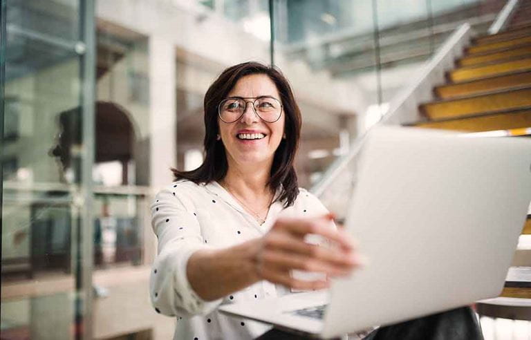 Elderly business professional standing and holding a laptop computer while smiling.