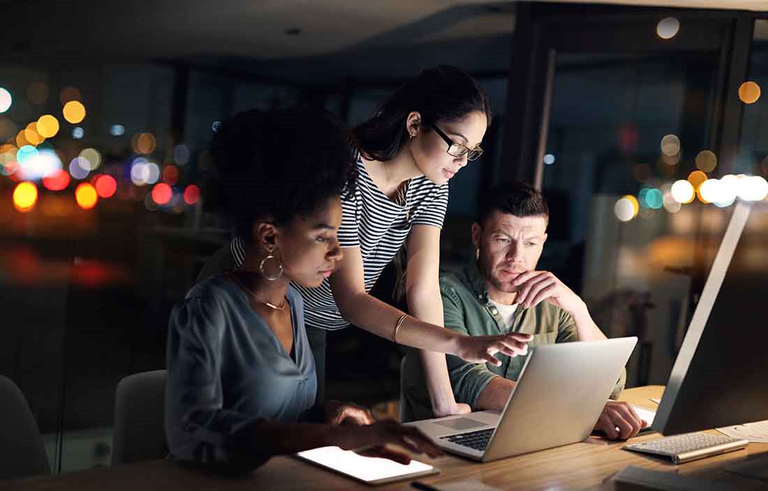 Three business professionals huddled around a laptop computer at night time reviewing information together.