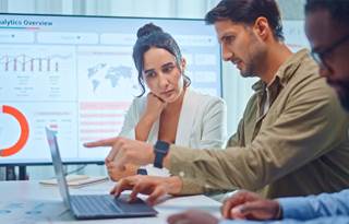 Three people in an office, looking at a laptop. Background shows charts