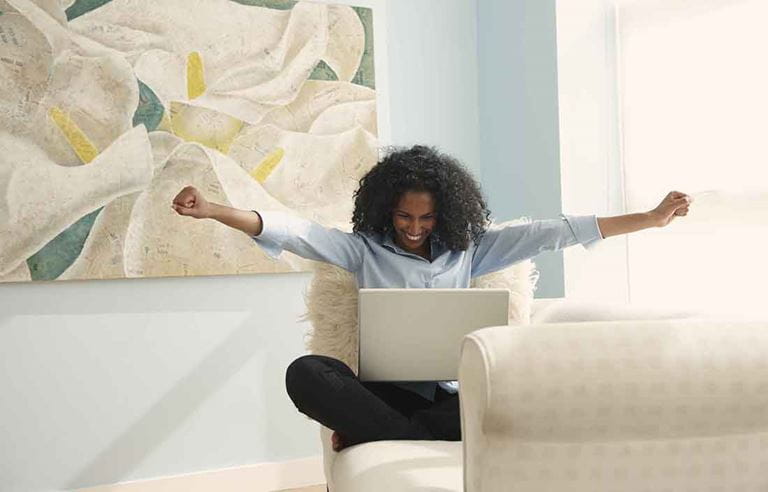 A woman sitting on a couch celebrating reading news off a laptop computer.