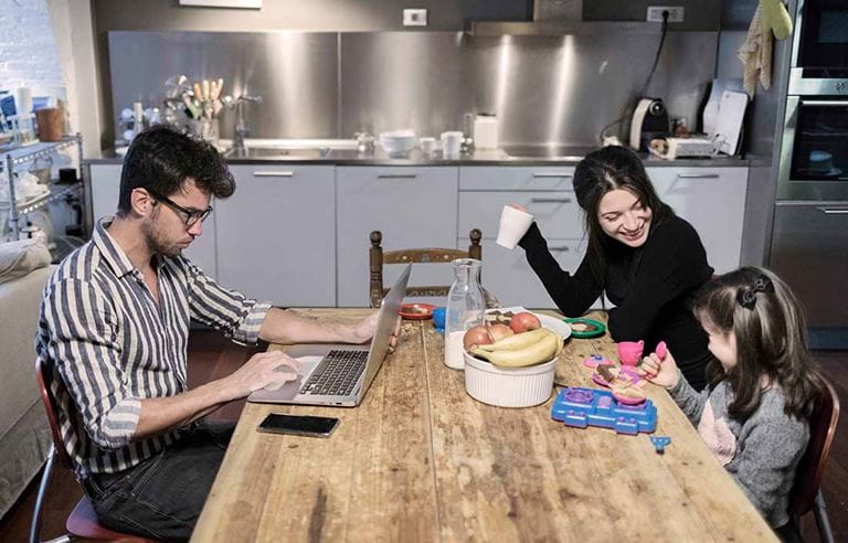 Parent working and sitting across from their family at the breakfast table.