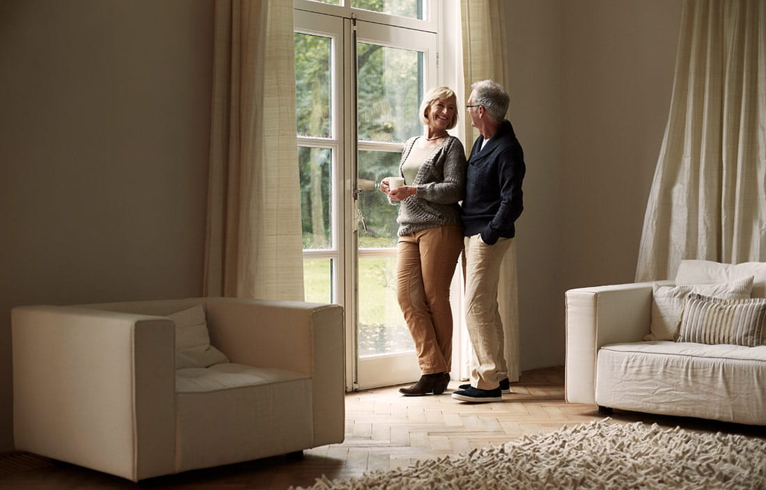 Male and female senior couple holding each other next to the window inside their home