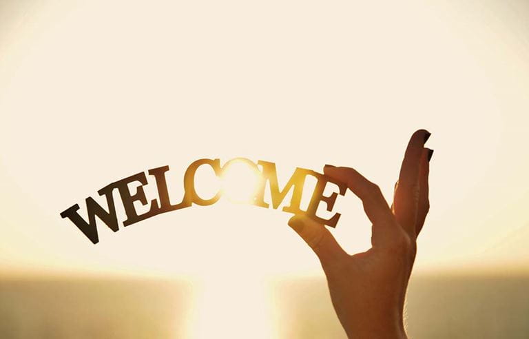 Hand holding up a wooden sculpture that says "welcome" against a sunrise or sunset light