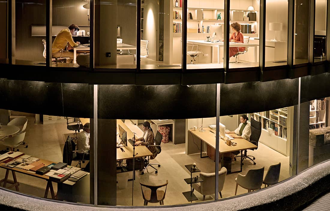 View from outside an office building looking in through the windows of two floors to see various people working at desks