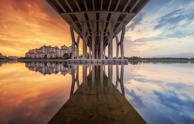 View under a bridge bisecting the horizon; on the left a city beyond the lake with an orange sky, on the right, blue sky