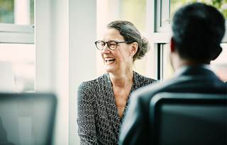 Woman in office setting laughing sitting across from a person at her desk.